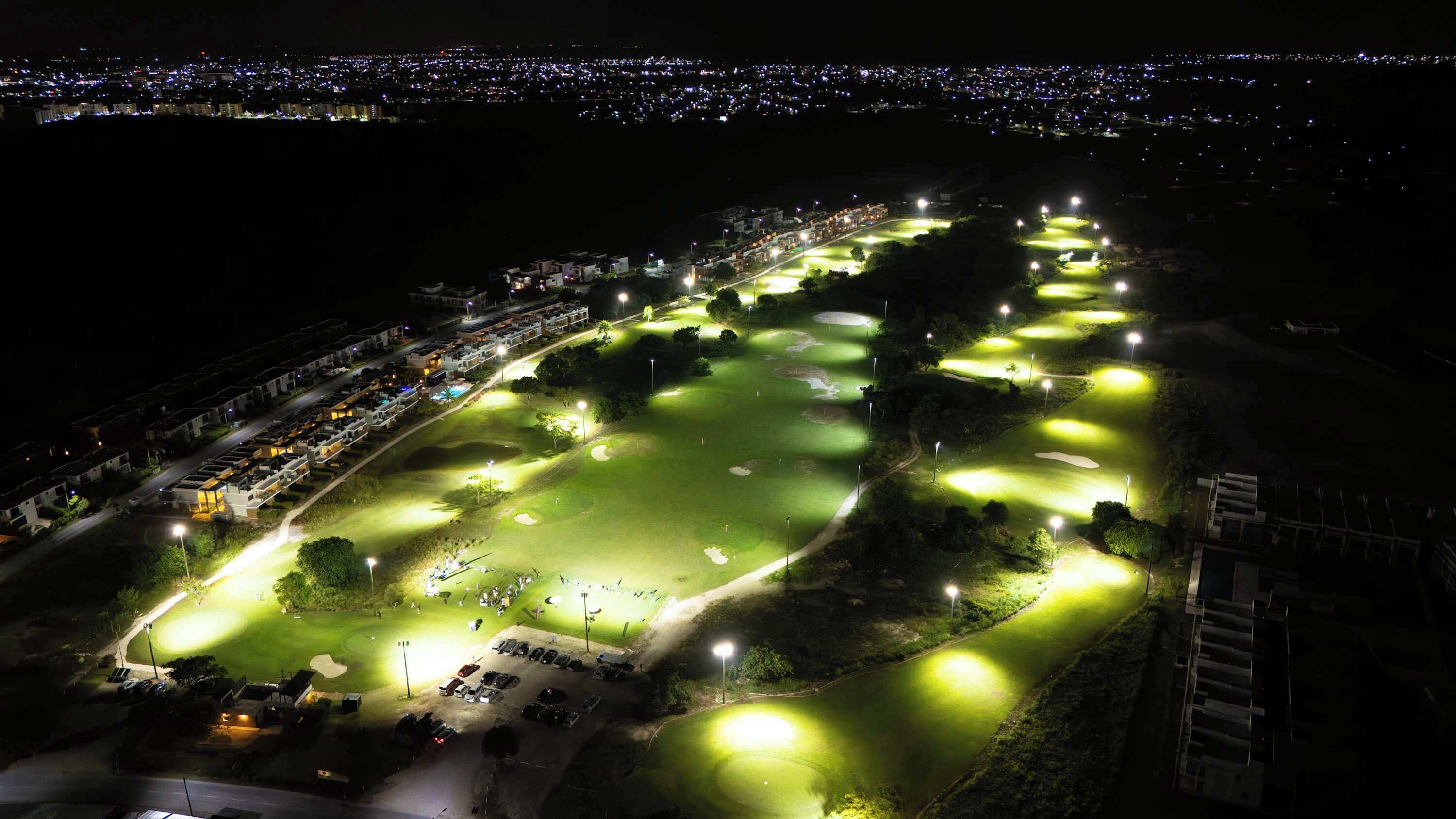 Een golfbaan 's nachts met verlichte fairways en greens, omgeven door woongebouwen.