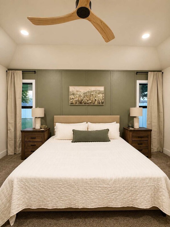 A bedroom with a large bed, two bedside tables, a ceiling fan, and a window with curtains.