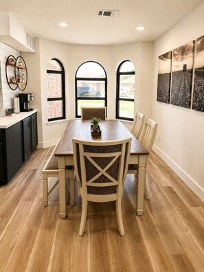 A dining area with a wooden table and chairs, a bench, and a kitchen area with cabinets and a countertop. - Rave On Retreat - 3BR/2BA - Family Friendly (Lubbock)