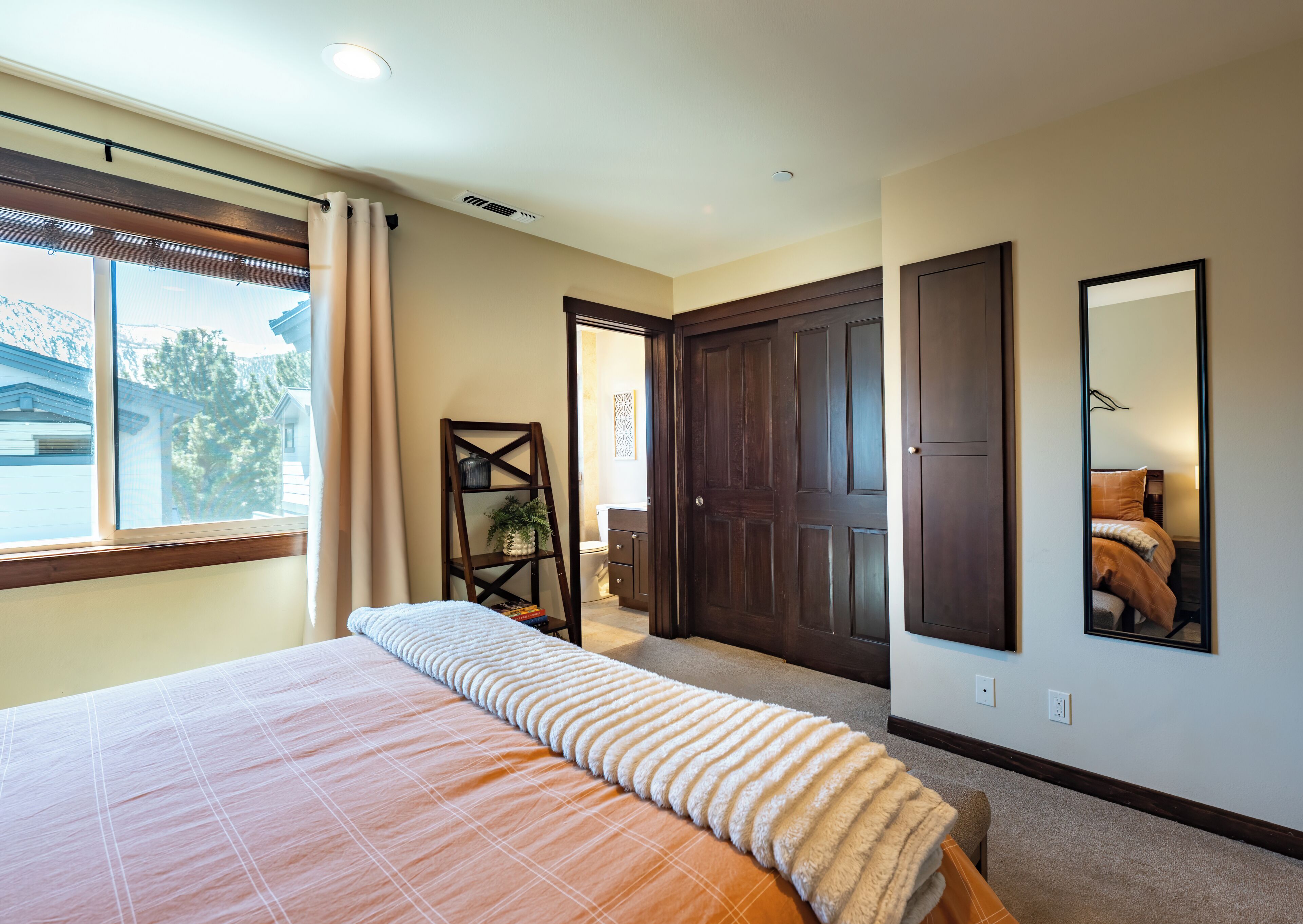 A bedroom with a large bed, a window with curtains, a wooden ladder shelf, and a dark wooden door.