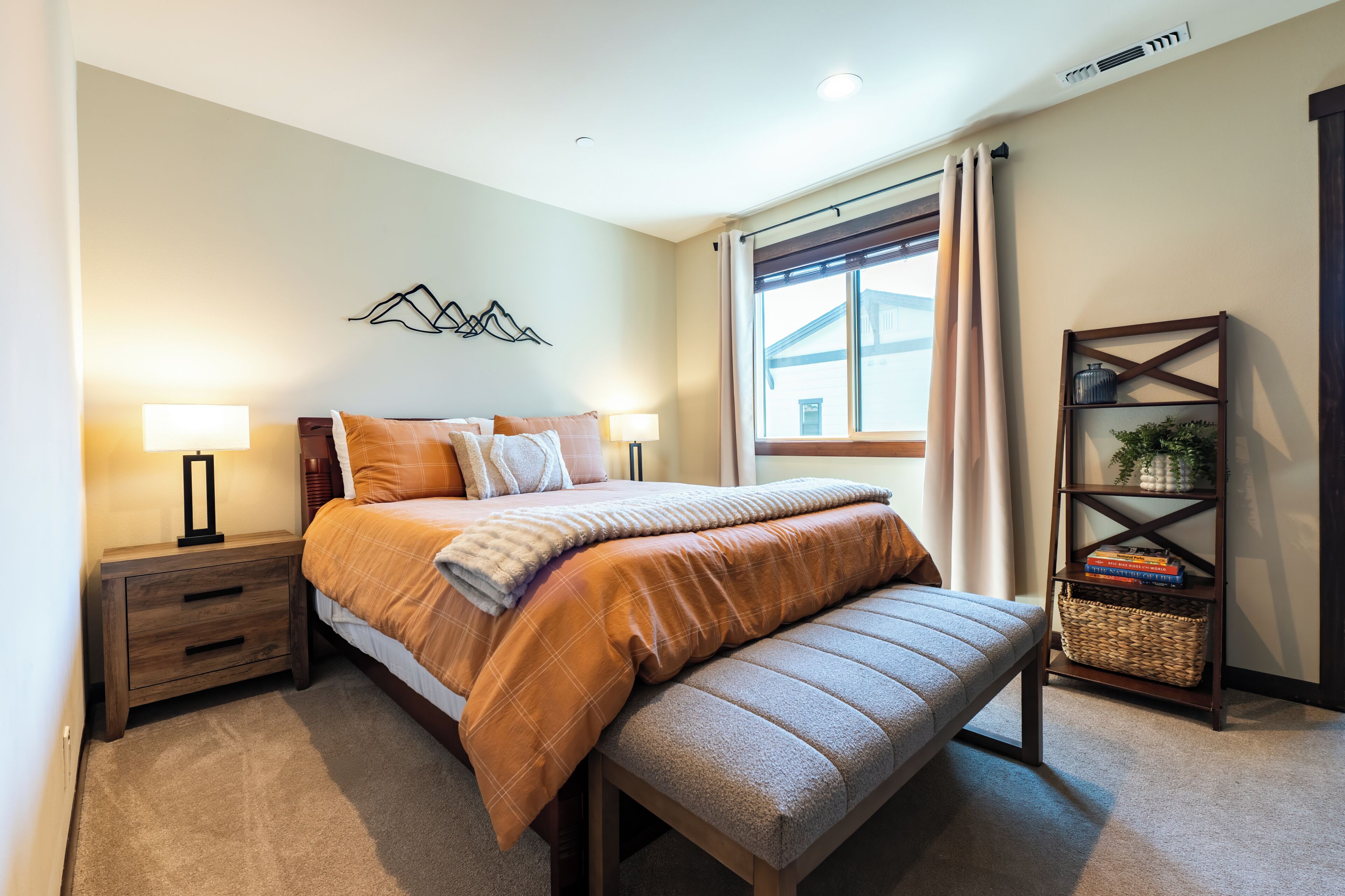 A bedroom with a bed, bedside tables, a bench, a wall-mounted shelf, and a window with curtains.
