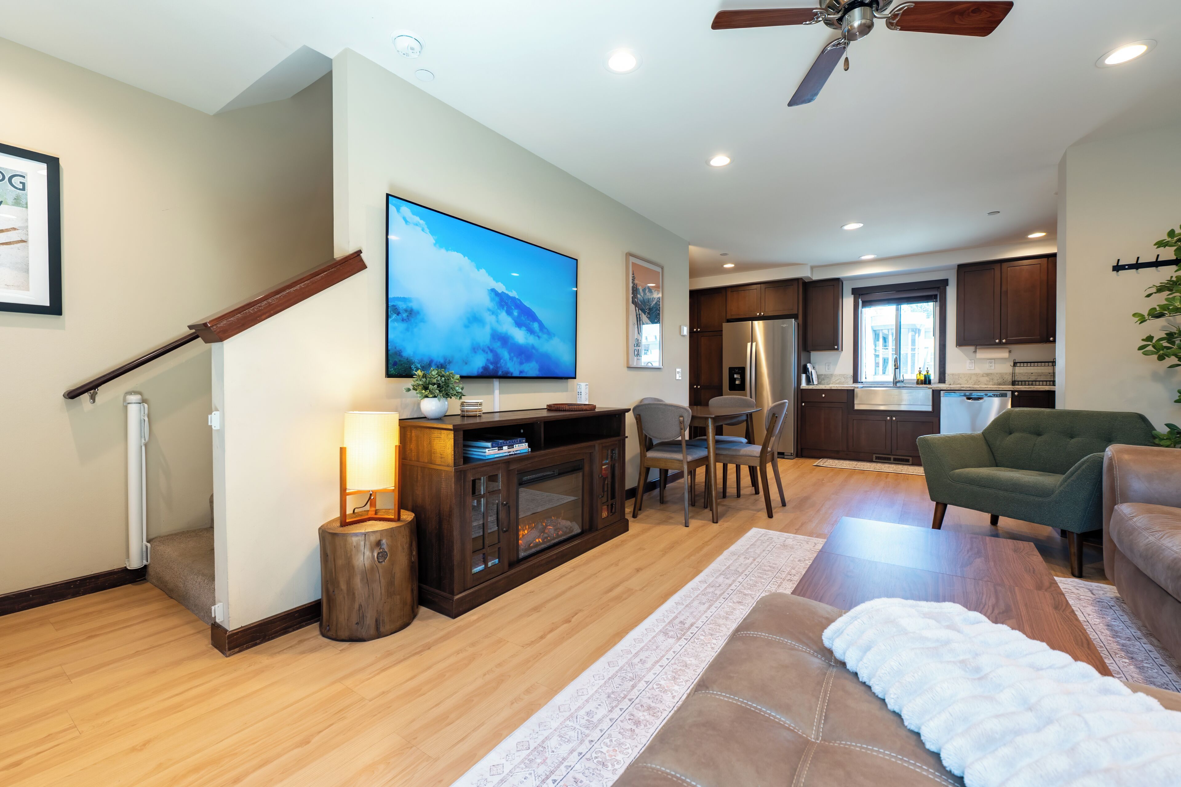 A modern living room with a flat-screen TV, wooden flooring, a dining area with chairs, a kitchen with dark cabinets, and a ceiling fan.