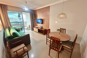 A modern living room with a sofa, a dining table with chairs, a TV on the wall, and a ceiling fan.