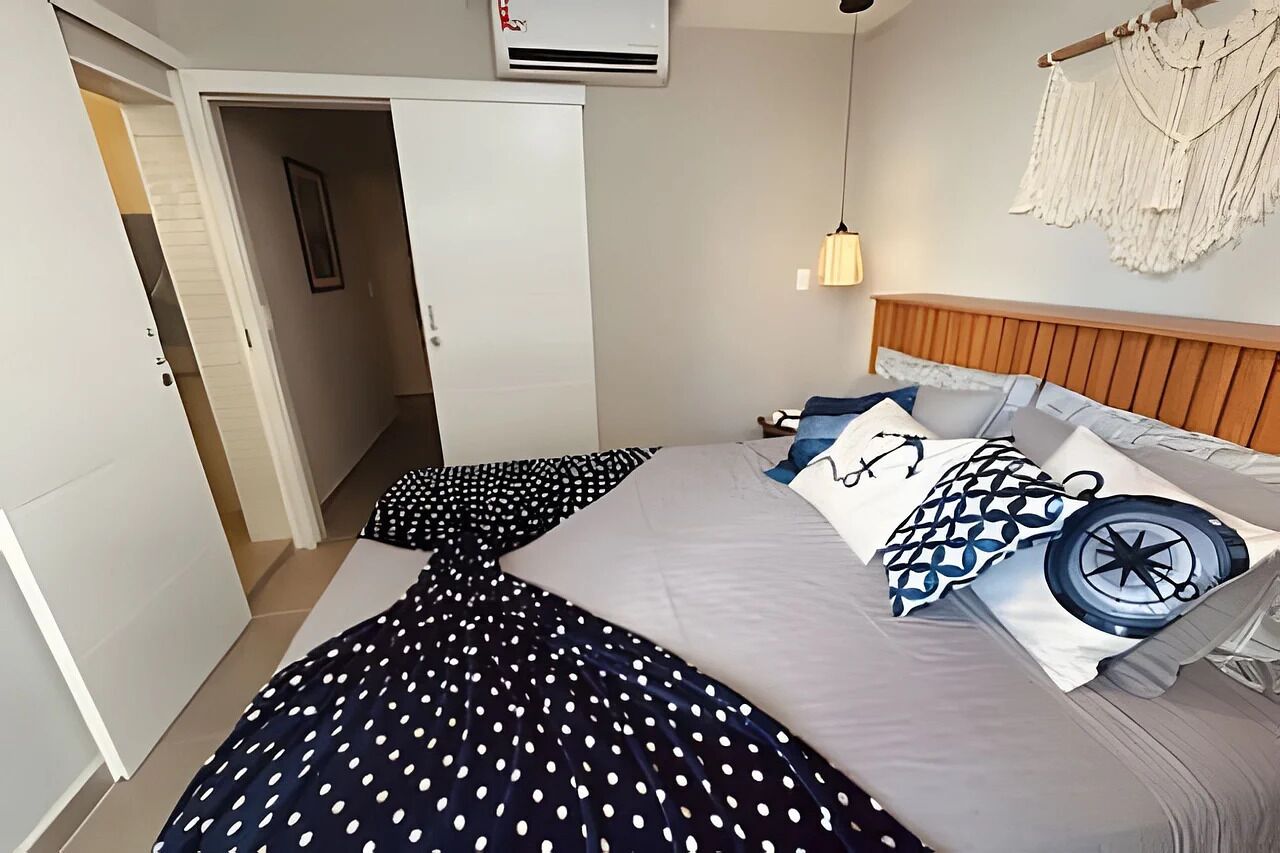 A bedroom with a bed, a wall-mounted air conditioner, a macrame wall hanging, and a door leading to another room.