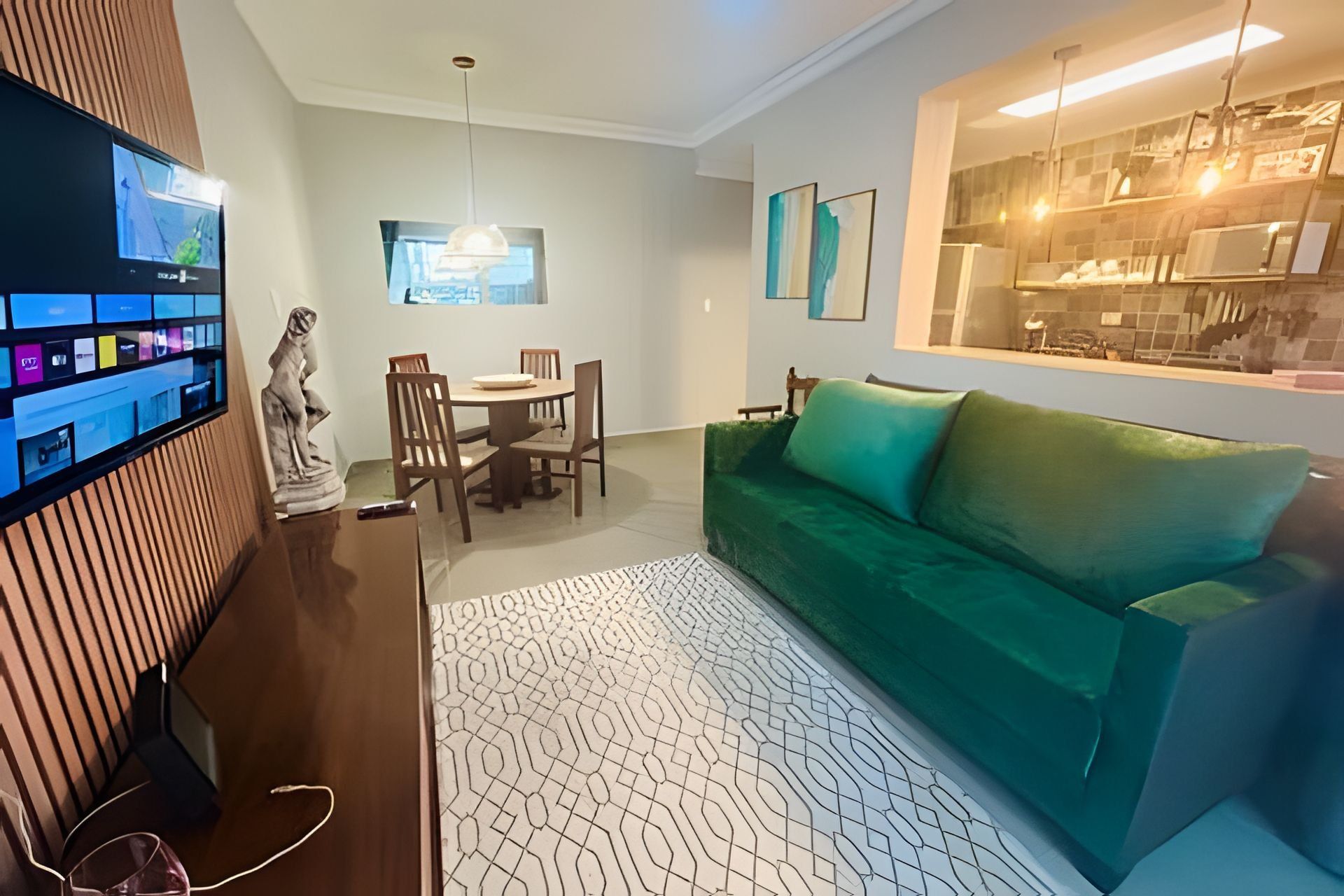 Een moderne woonkamer met een groene bank, een eettafel met stoelen, een televisie bevestigd aan een houten paneel, en een zichtbare keukenruimte via een glazen scheiding.