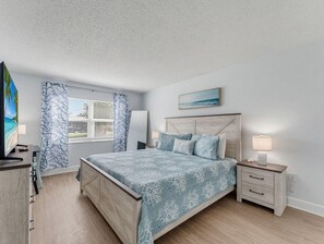 A bedroom with a bed, a nightstand, a TV, and a window with curtains. - El Mar Condos #113 - "three Sisters" (North Redington Beach)