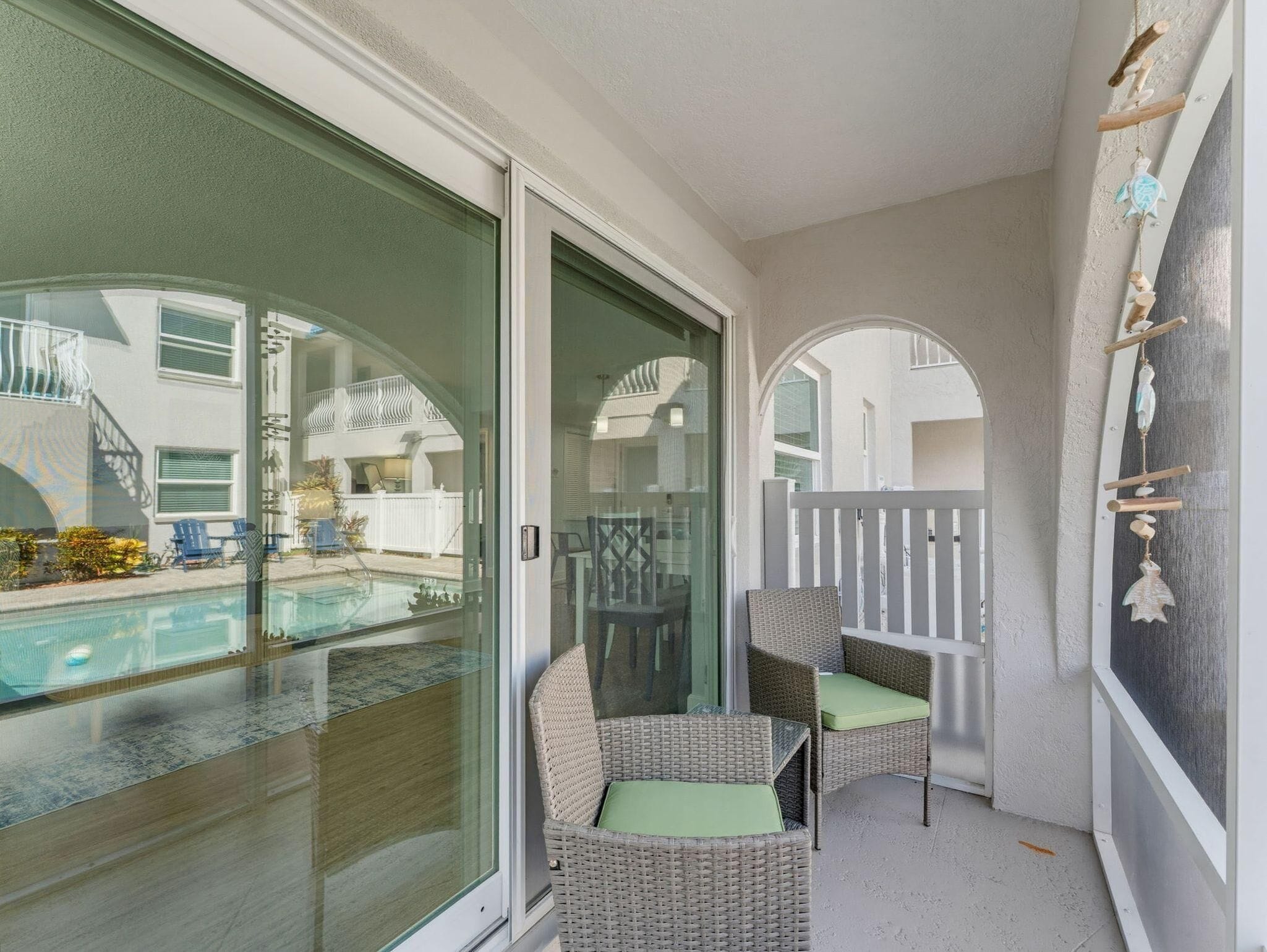 A balcony with a glass door, wicker chairs, a small pool, and a view of a house with a white fence.