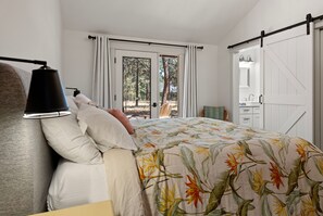 Ein Schlafzimmer mit einem Bett, einem Nachttisch, einer Lampe, einem Fenster mit Vorhängen und Blick auf Bäume.