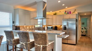 A modern kitchen with white cabinets, a stainless steel refrigerator, a built-in microwave, and a granite countertop. There are three woven chairs at the island. - Waterfront, Heated-chilled pool, Outdoor kitchen, downstairs bathroom w/ shower! (cudjoe key)