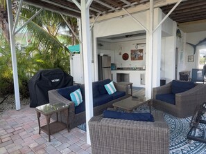 A covered outdoor seating area with wicker furniture and a grill, leading to a kitchen area. - Waterfront, Heated-chilled pool, Outdoor kitchen, downstairs bathroom w/ shower! (cudjoe key)
