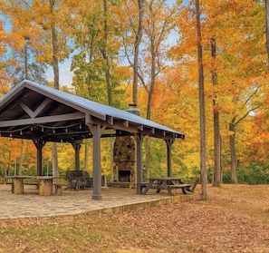 A covered picnic area with benches and a stone fireplace, surrounded by trees with autumn foliage. - Everly House | Waterfall + Private Trails (Monteagle)