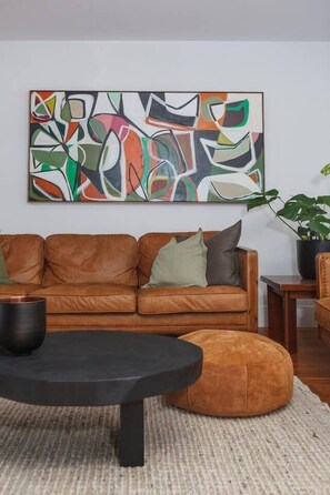 Ein Wohnzimmer mit einem braunen Ledersofa, einem runden schwarzen Couchtisch, einem runden Hocker und einem großen abstrakten Gemälde an der Wand.