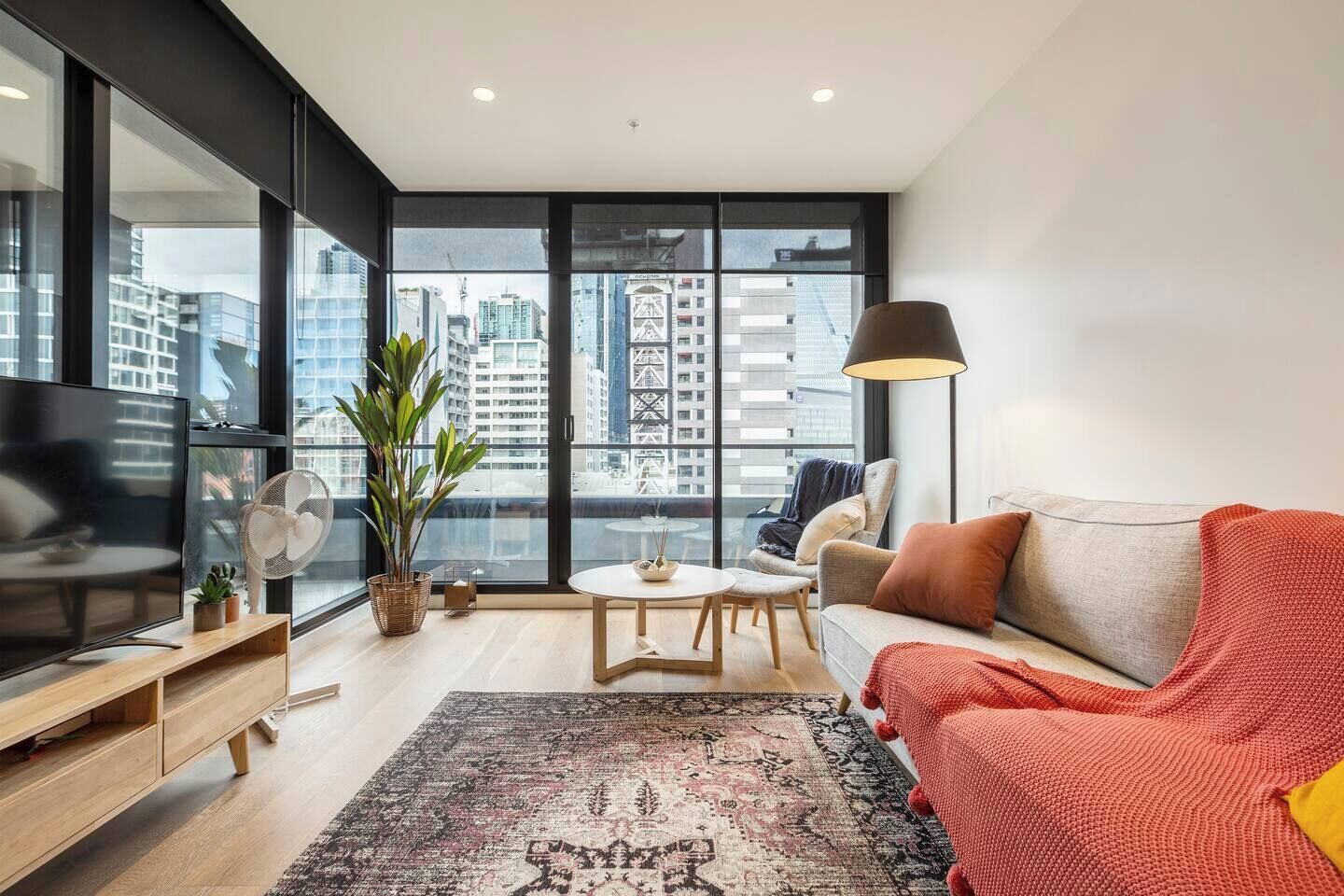 A modern living room with a large window, a sofa, a TV, a fan, and a table with a plant.
