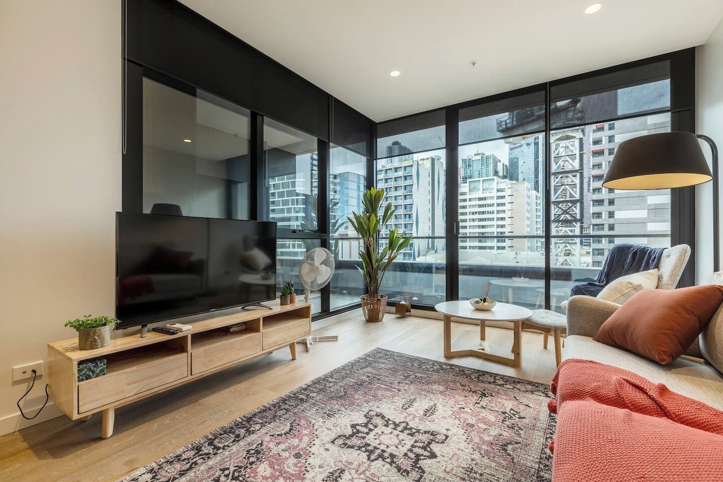 Una sala de estar moderna con un televisor grande, un mueble de entretenimiento de madera, una mesa redonda de centro, un sofá y vistas a un paisaje urbano a través de amplios ventanales.