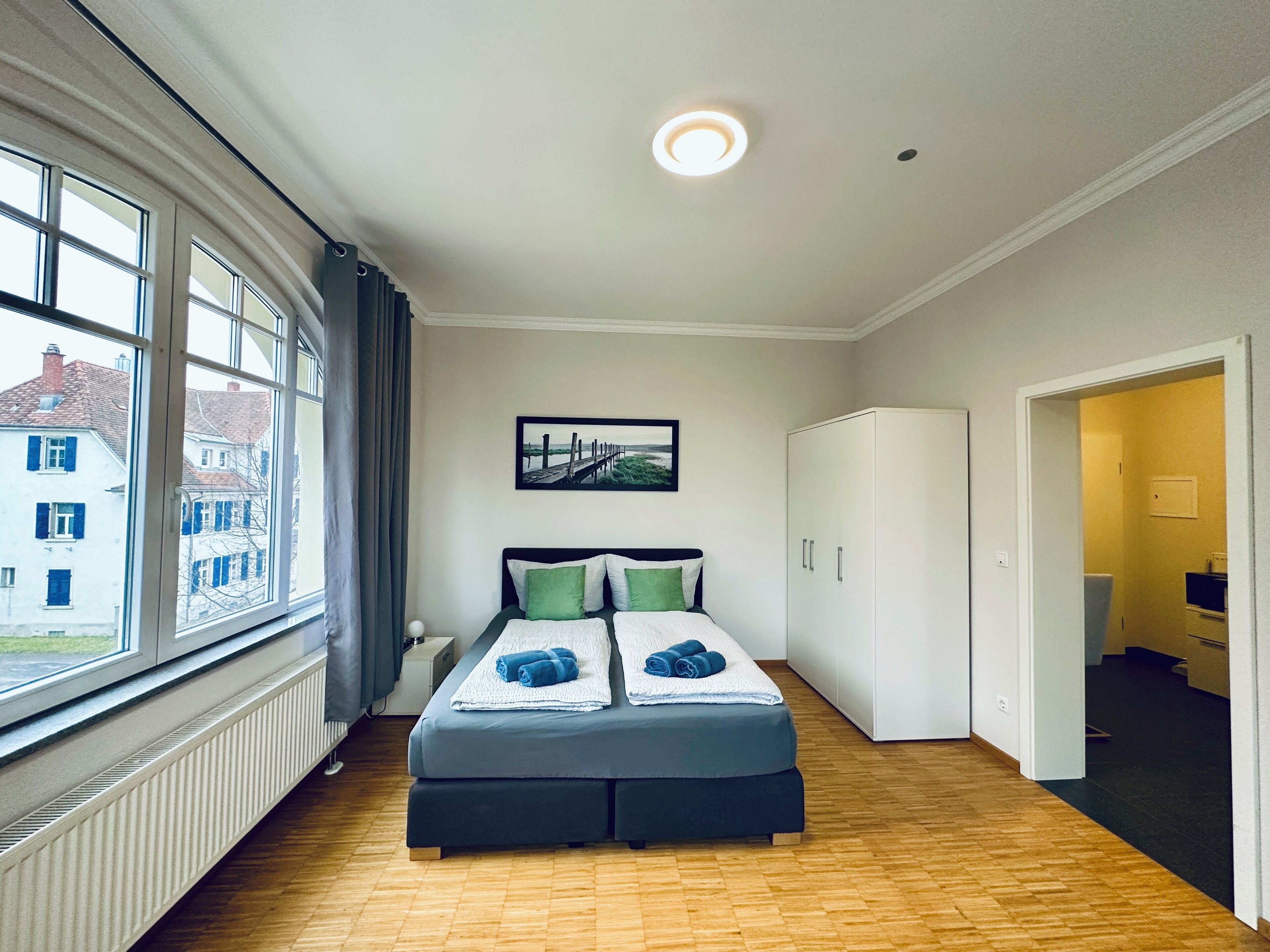 Ein Hotelzimmer mit großem Fenster, einem Bett mit Kissen, einem Heizkörper und einer Tür zu einem Badezimmer.