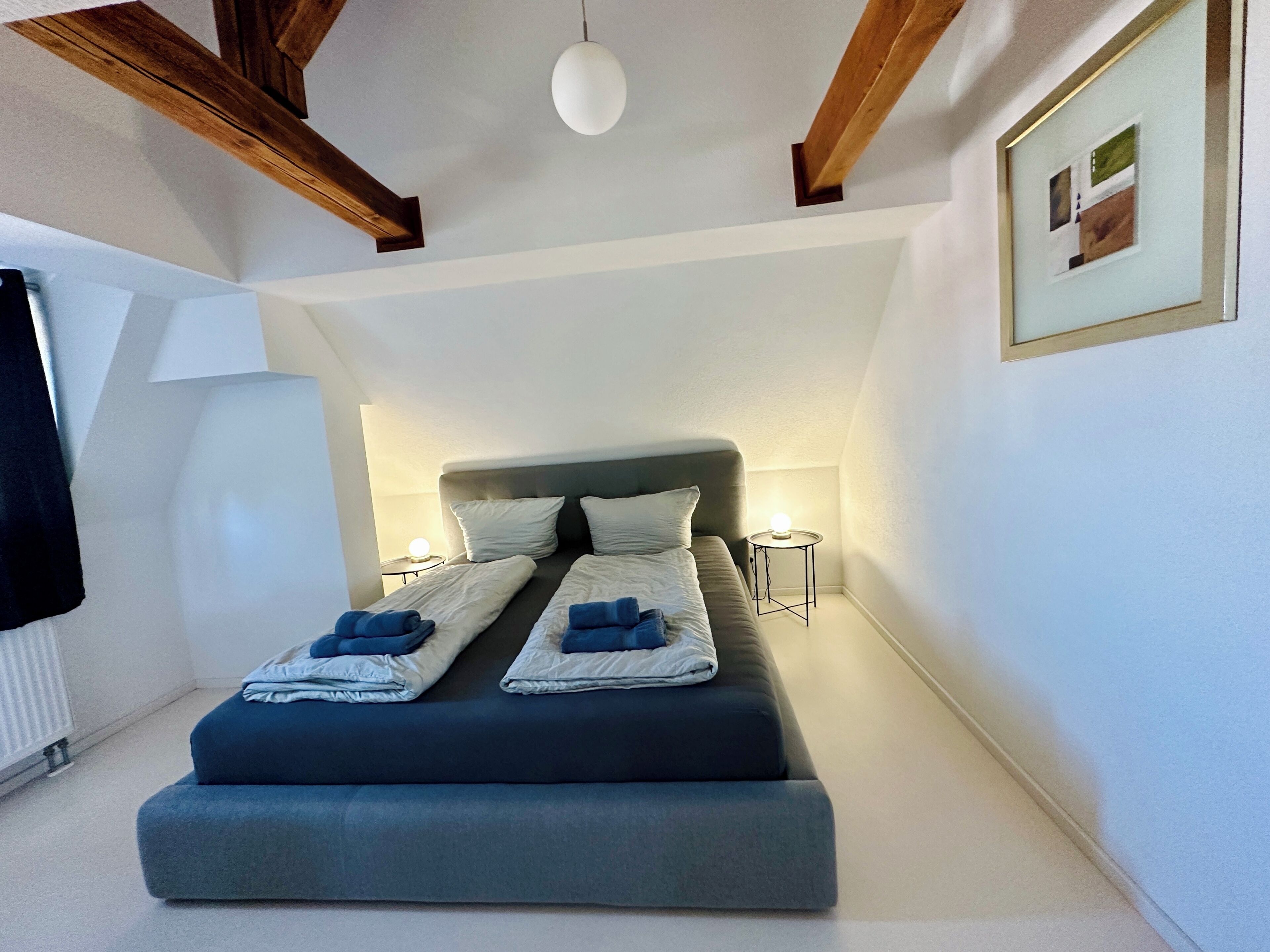 Ein Schlafzimmer mit einem Bett, Nachttischen, einer Pendelleuchte und einem gerahmten Bild an der Wand.