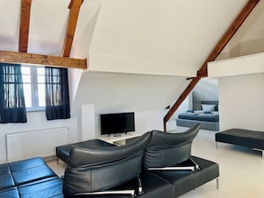 Un dormitorio tipo loft moderno con techo inclinado, un sofá de cuero negro, un televisor de pantalla plana y una cama con ropa de cama azul.