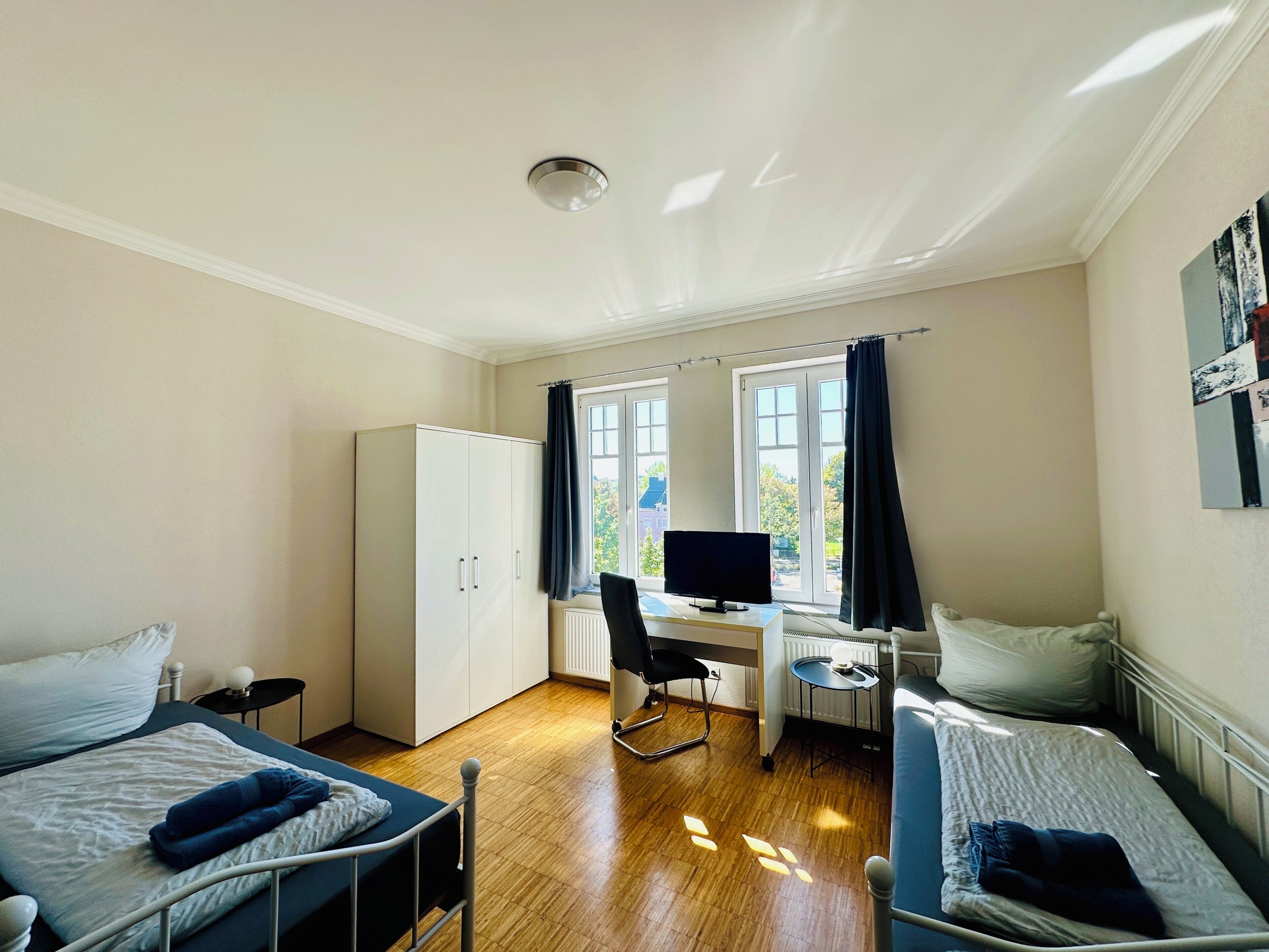 Una habitación con dos camas, un escritorio con computadora, un armario y una ventana con cortinas.
