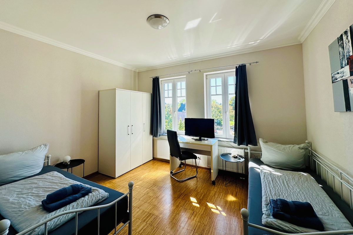 A room with two beds, a desk with a computer, a wardrobe, and a window with curtains.