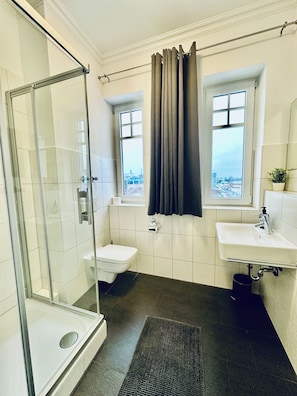 A modern bathroom with a glass shower enclosure, a toilet, a sink, and a window with curtains. - Apartmenthouse Singen  (Singen (Hohentwiel))