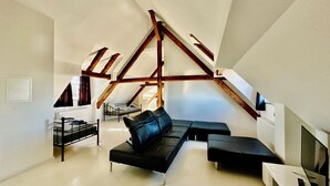 A modern loft bedroom with a sloped ceiling, a black leather sofa, a bed, and a television.