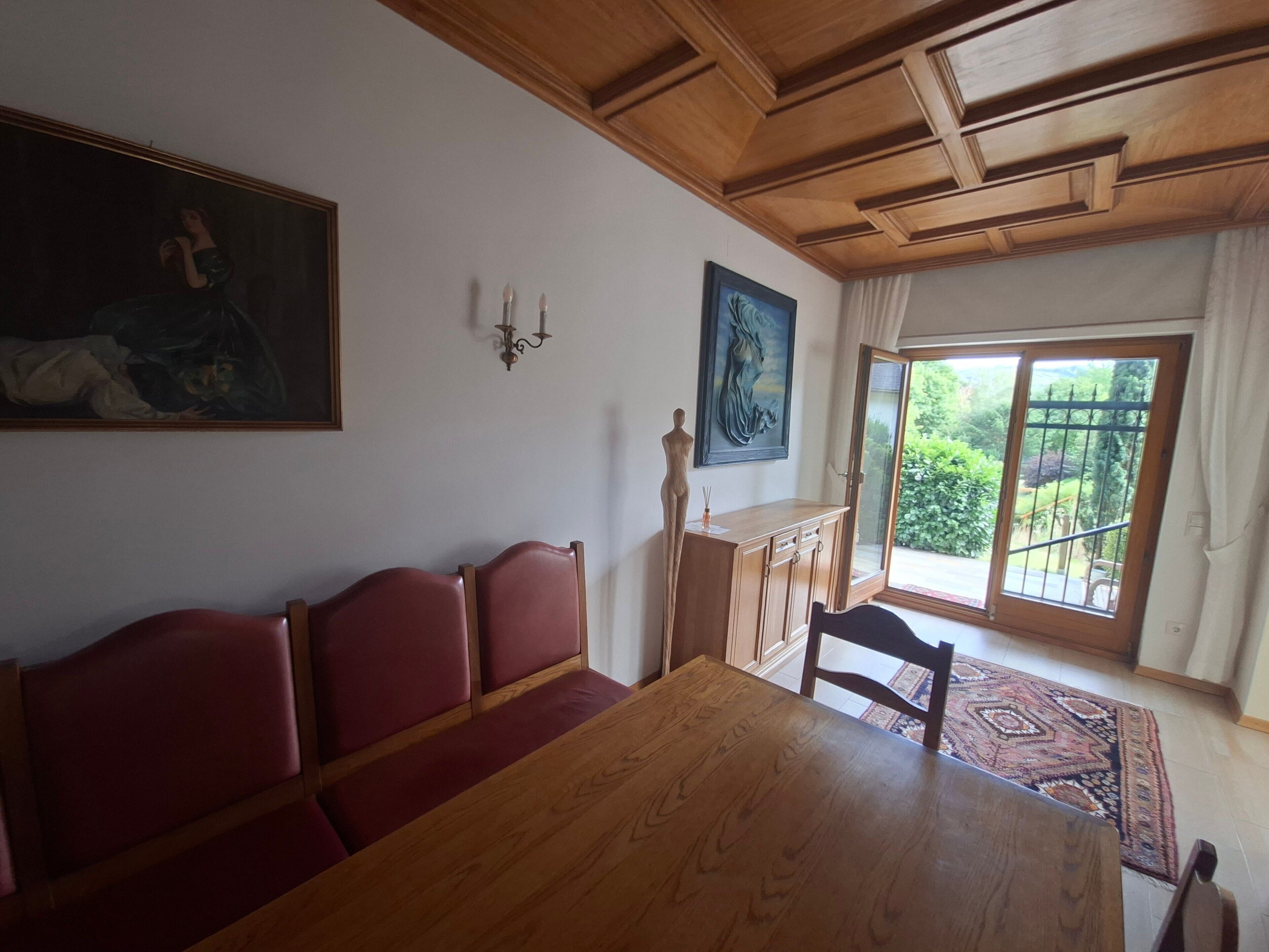 Ein Essbereich mit einem Holztisch, roten Polsterstühlen, einem Gemälde an der Wand und Blick durch eine Glastür.