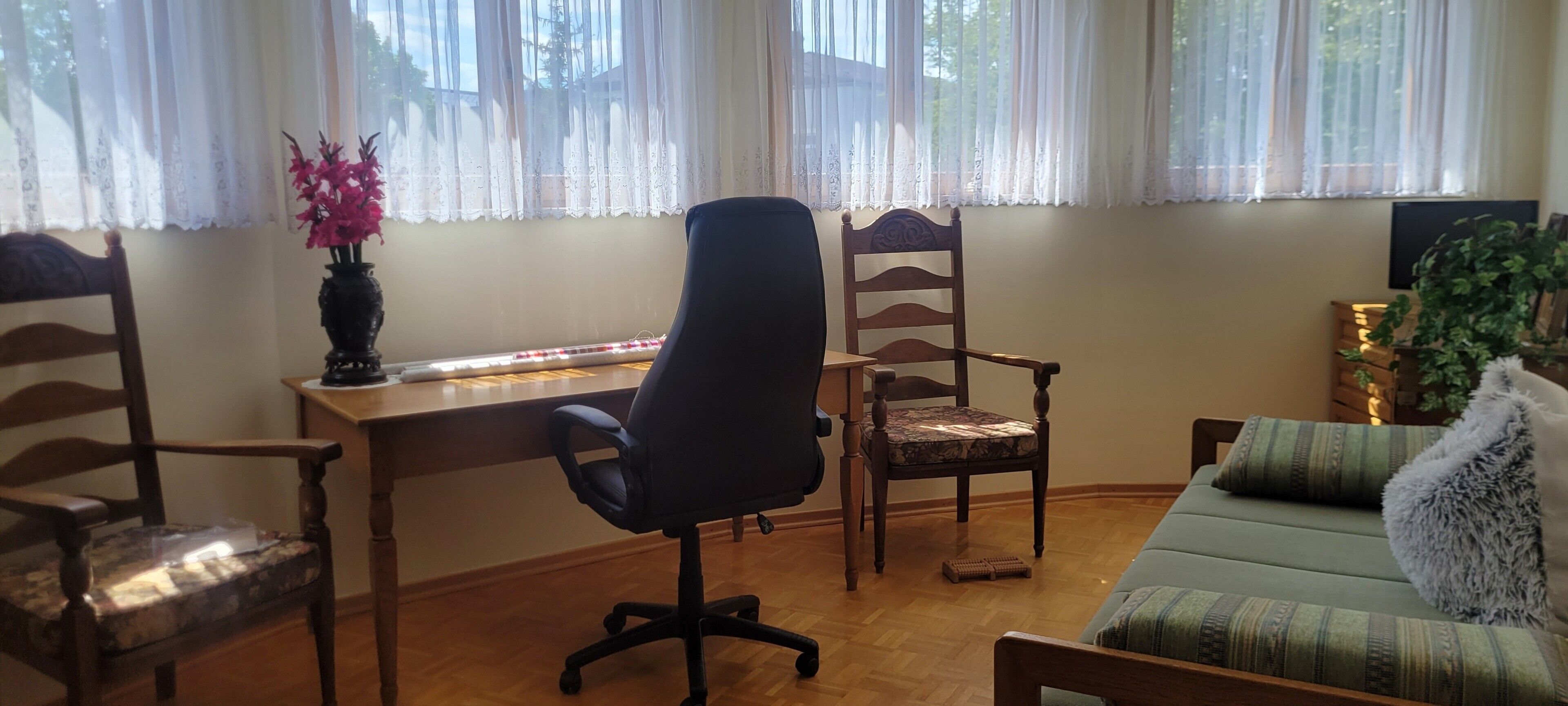 A room with a wooden desk, two chairs, a sofa, and a window with sheer curtains.