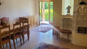 Uma área de refeições com cadeiras de madeira e uma mesa, um tapete estampado e vista para um espaço ao ar livre verde através de uma porta aberta.