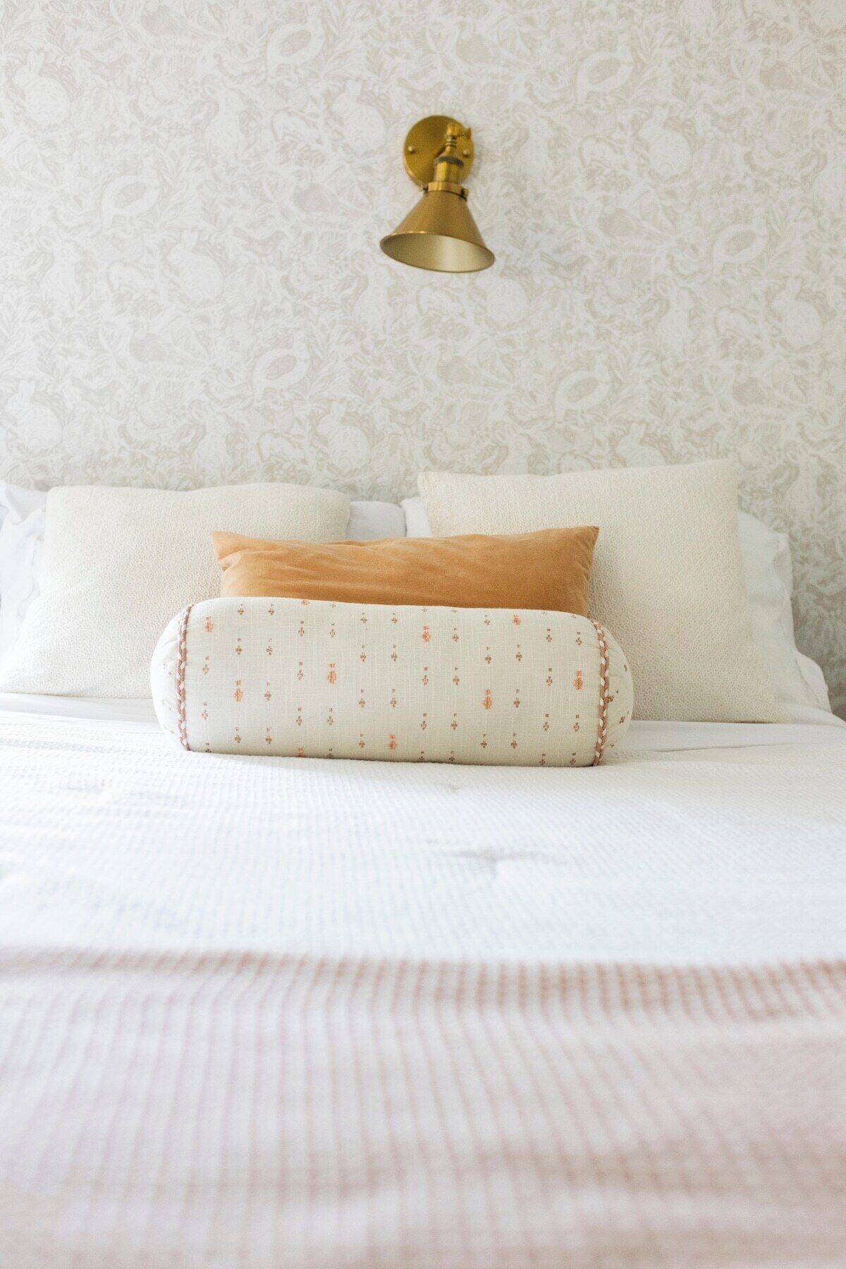 A bed with white bedding and a patterned pillow, a wall-mounted light fixture, and a patterned wallpaper.