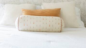 A bed with white bedding and a patterned pillow, a wall-mounted light fixture, and a patterned wallpaper.