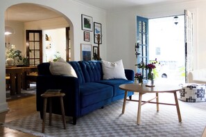 Una sala de estar con un sofá azul, una mesa de centro redonda de madera y una alfombra a cuadros. Se observa una puerta que conduce a otra habitación, y un pequeño taburete.
