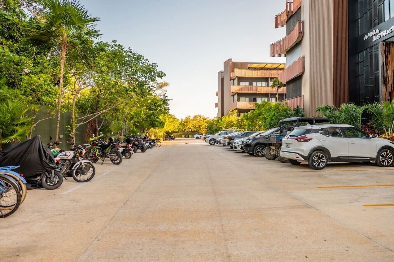 Un espace de stationnement où sont garées de nombreuses voitures et motos, entouré d’arbres et d’immeubles.