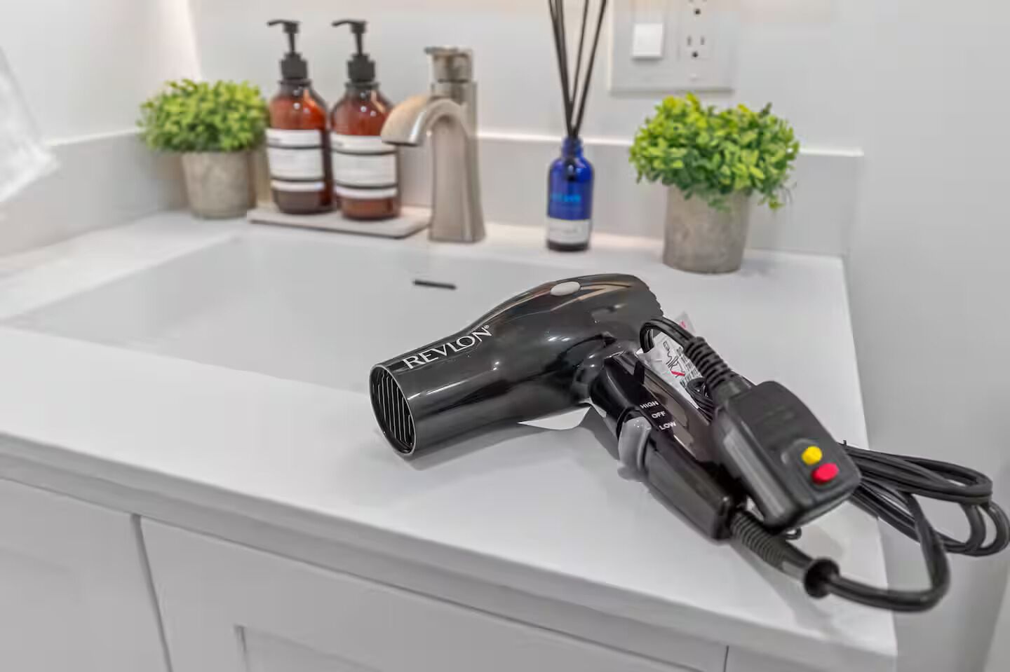 Een haardroger op een wit aanrecht met een wastafel, een kraan en twee potplanten.