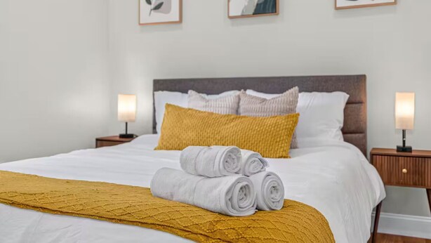 Una cama bien tendida con edredón blanco, una manta color mostaza y toallas enrolladas. Hay dos mesitas de noche con lámparas, y las paredes están decoradas con obras de arte enmarcadas.