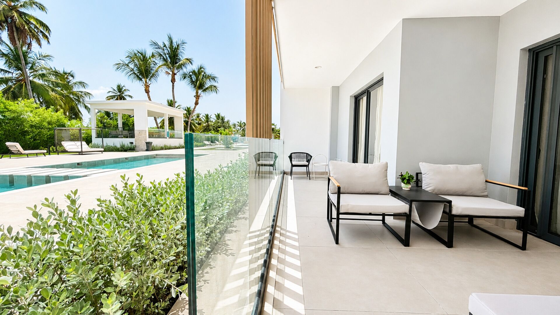 A modern outdoor balcony with a pool, lounge chairs, and a view of a resort area.