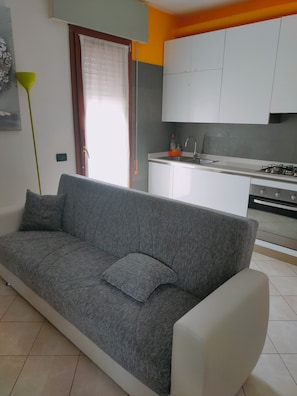 A modern living room with a grey sofa, a small kitchen area with white cabinets, and a window with a curtain. - LA BELLA Flat, Super Easy Venice, Bus stop right in front of Venice (short ride) (Venezia)