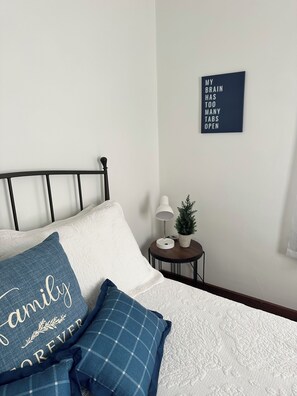 En pænt redt seng med et hvidt dynebetræk og to puder, hvoraf den ene har et blåt mønster og den anden et hvidt design. Et lille natbord med en lampe og en pyntende plante er synligt.