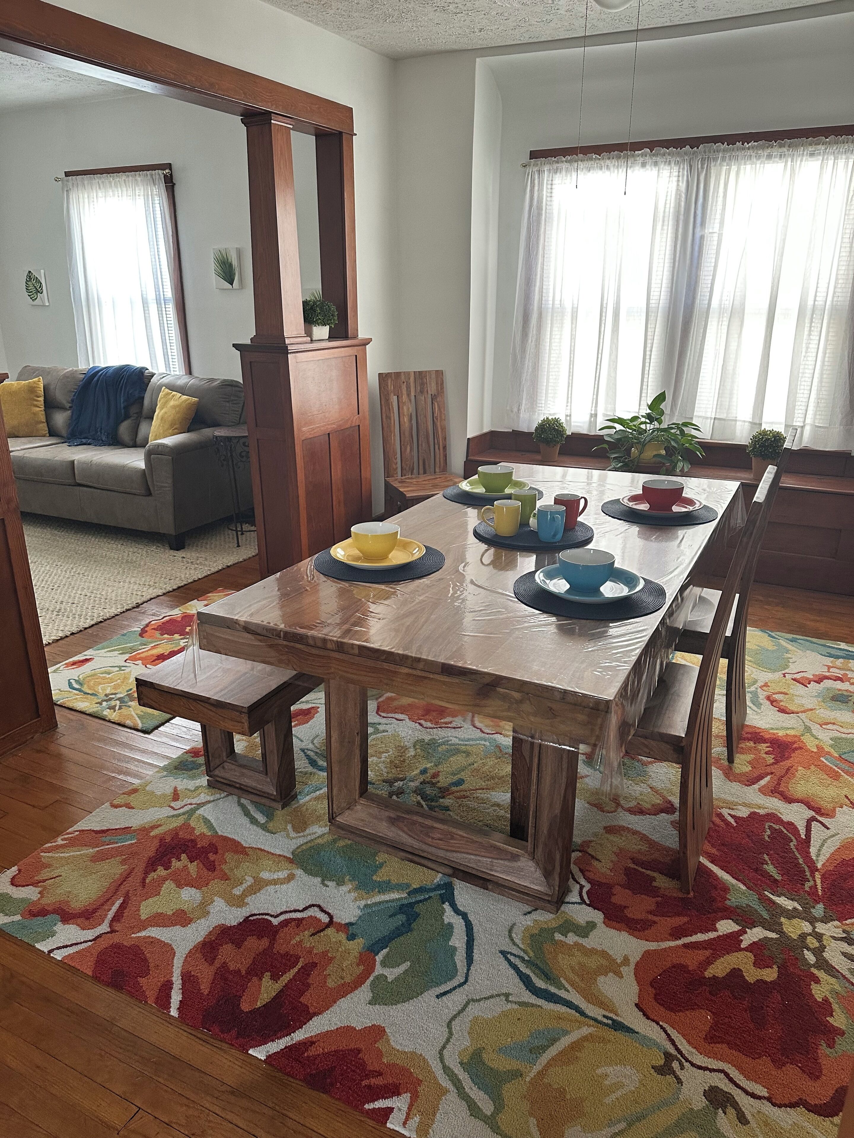 Un comedor con una mesa de madera dispuesta para cuatro personas, rodeada de sillas. Se ve un sofá y un banco de madera al fondo.