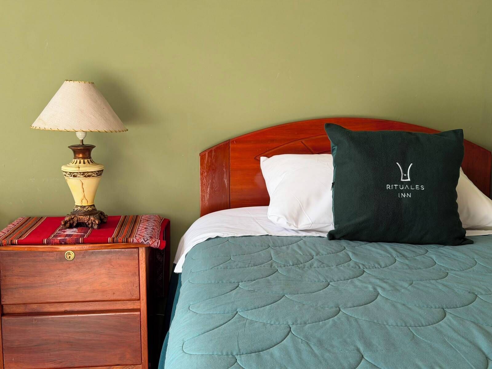 Una cama bien hecha con una colcha color turquesa, un almohadón blanco y un almohadón verde oscuro con un logotipo. Una mesita de noche de madera con una lámpara y un paño estampado está al lado de la cama.