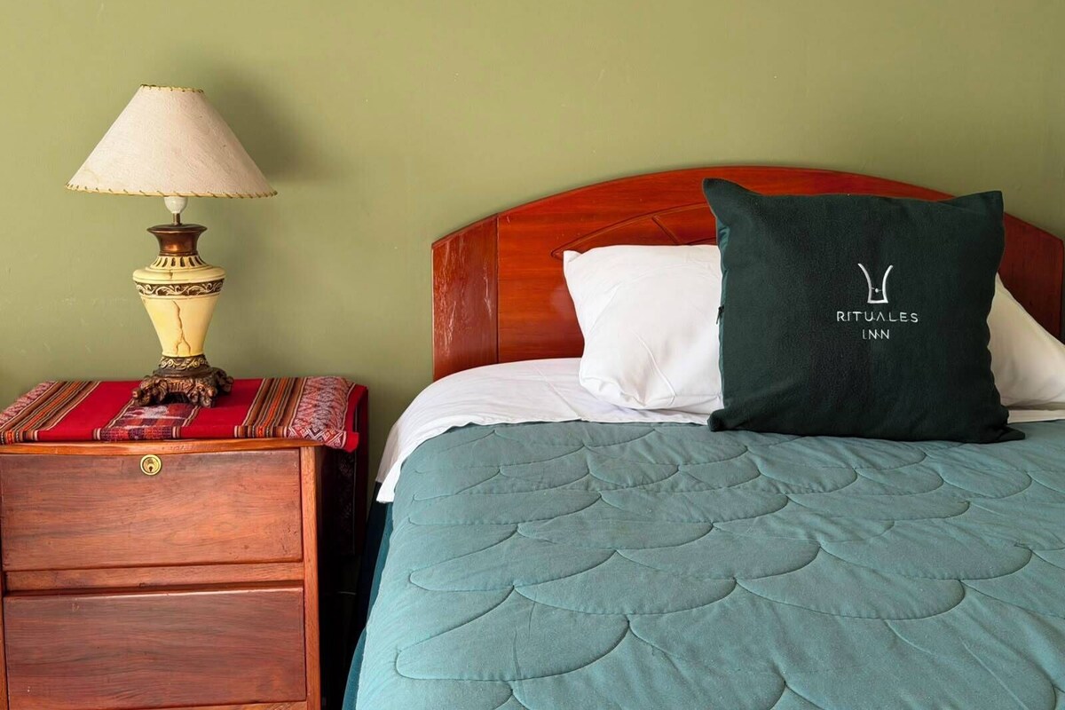 A neatly made bed with a teal quilt, a white pillow, and a dark green pillow with a logo. A wooden nightstand with a lamp and a patterned cloth is beside the bed.