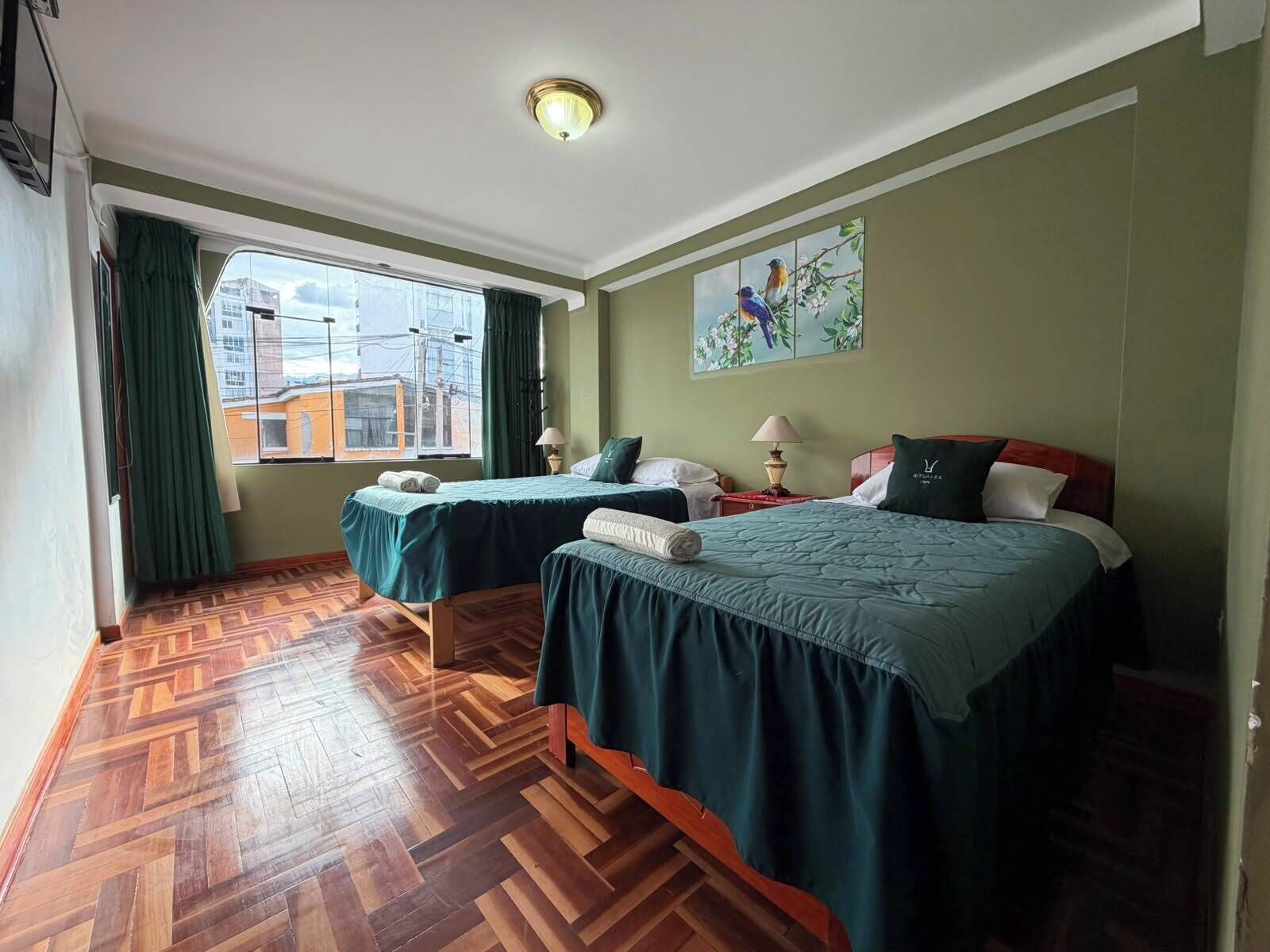 A hotel room with two beds, a large window, a wooden parquet floor, and a painting on the wall.