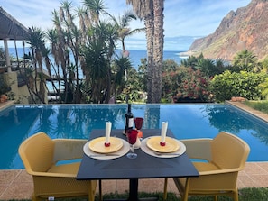 Un espace repas aménagé au bord de la piscine, avec une table dressée pour deux personnes, offrant une vue sur un paysage côtier agrémenté de palmiers et sur la mer.