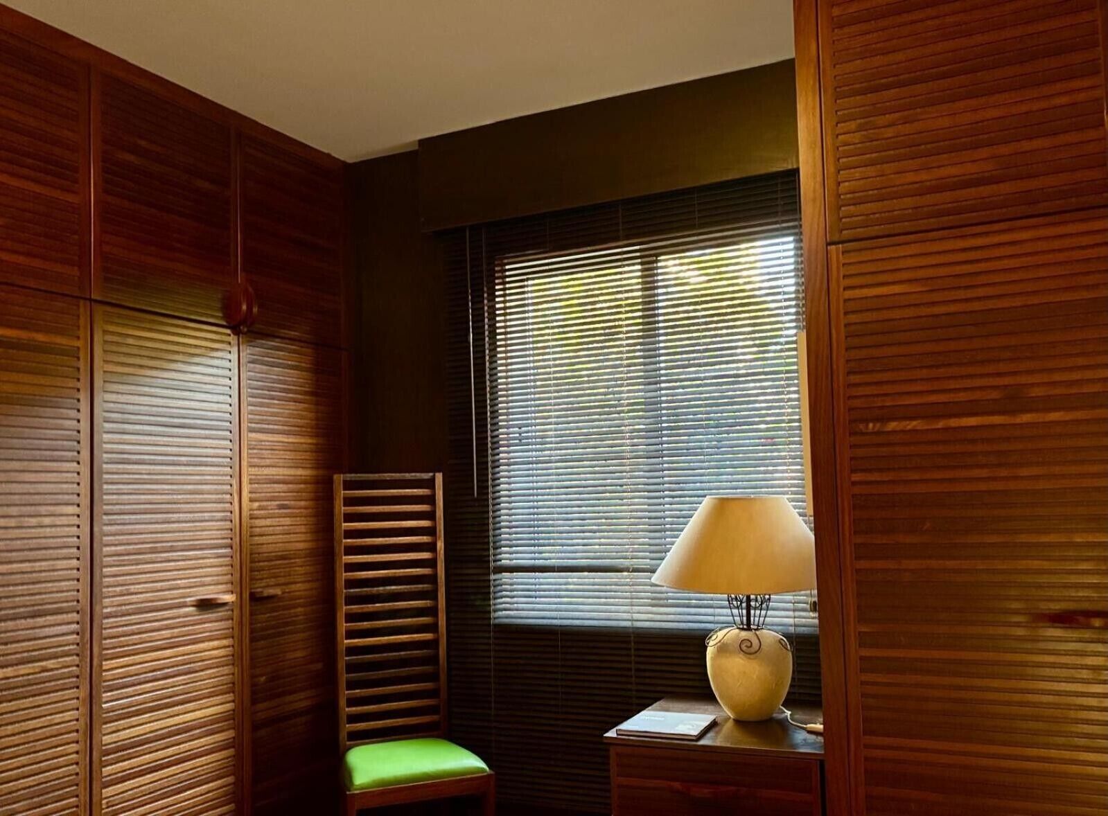 Una habitación con paneles de madera, una ventana con persianas, un sillón verde acolchado y una mesa con una lámpara.