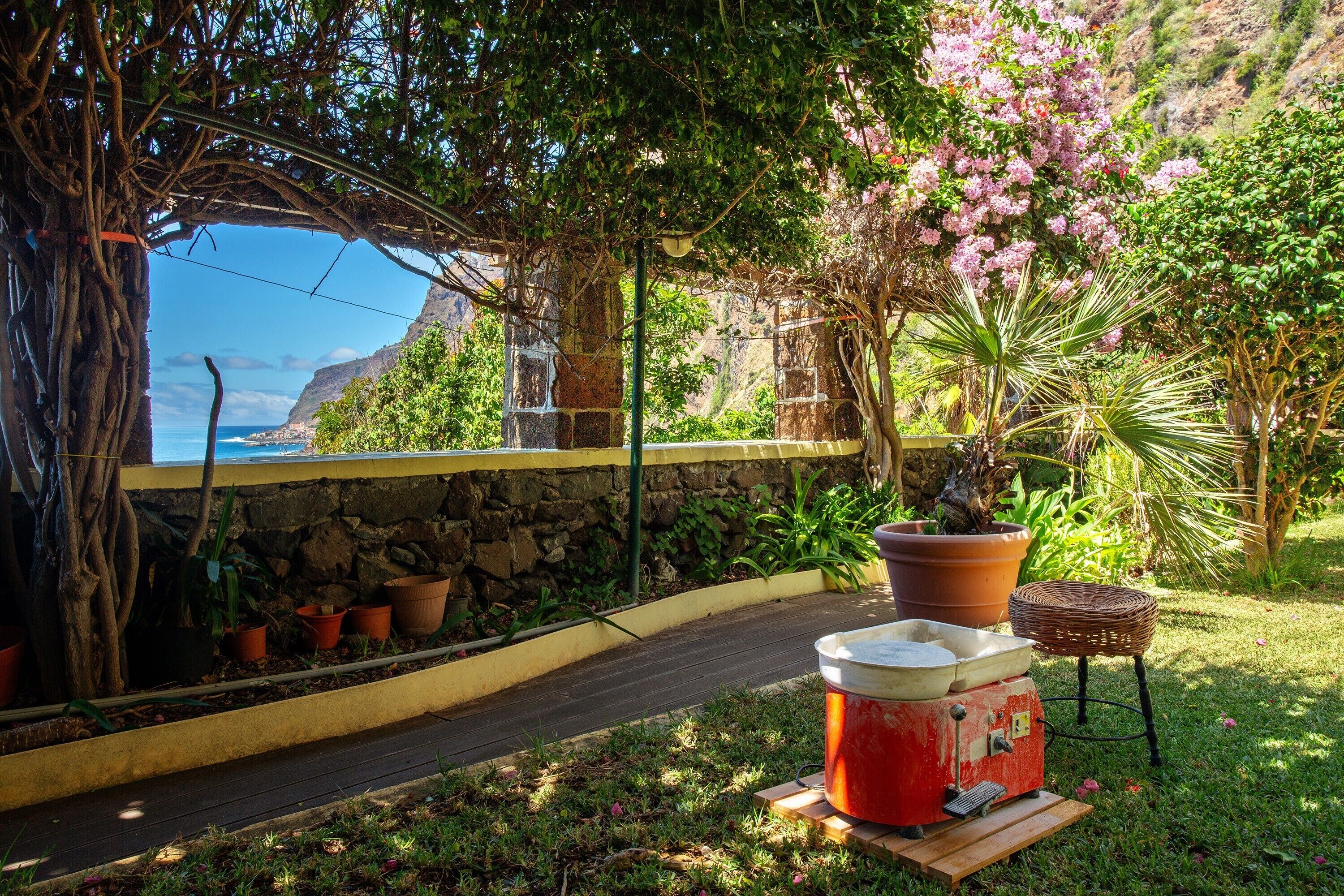 Un jardin avec un mur de pierres, des plantes en pot, une table en bois avec des bols, et une vue sur l’océan.