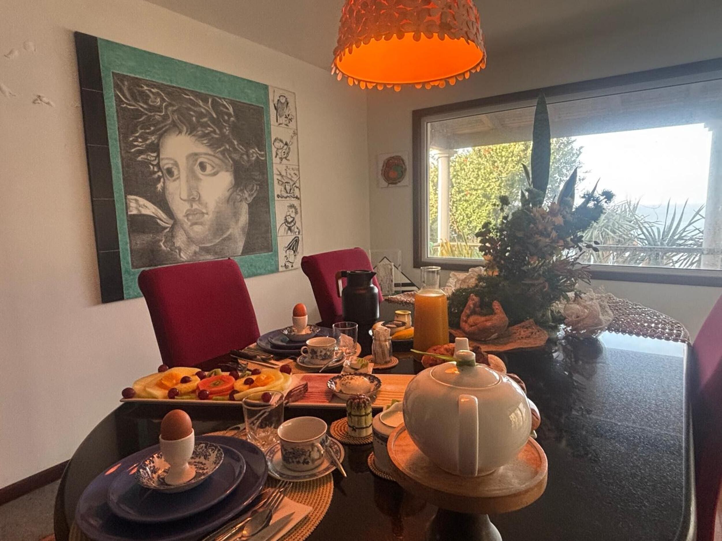 Una mesa de comedor dispuesta con platos, tazas y una tetera, con un cuadro y una ventana al fondo.