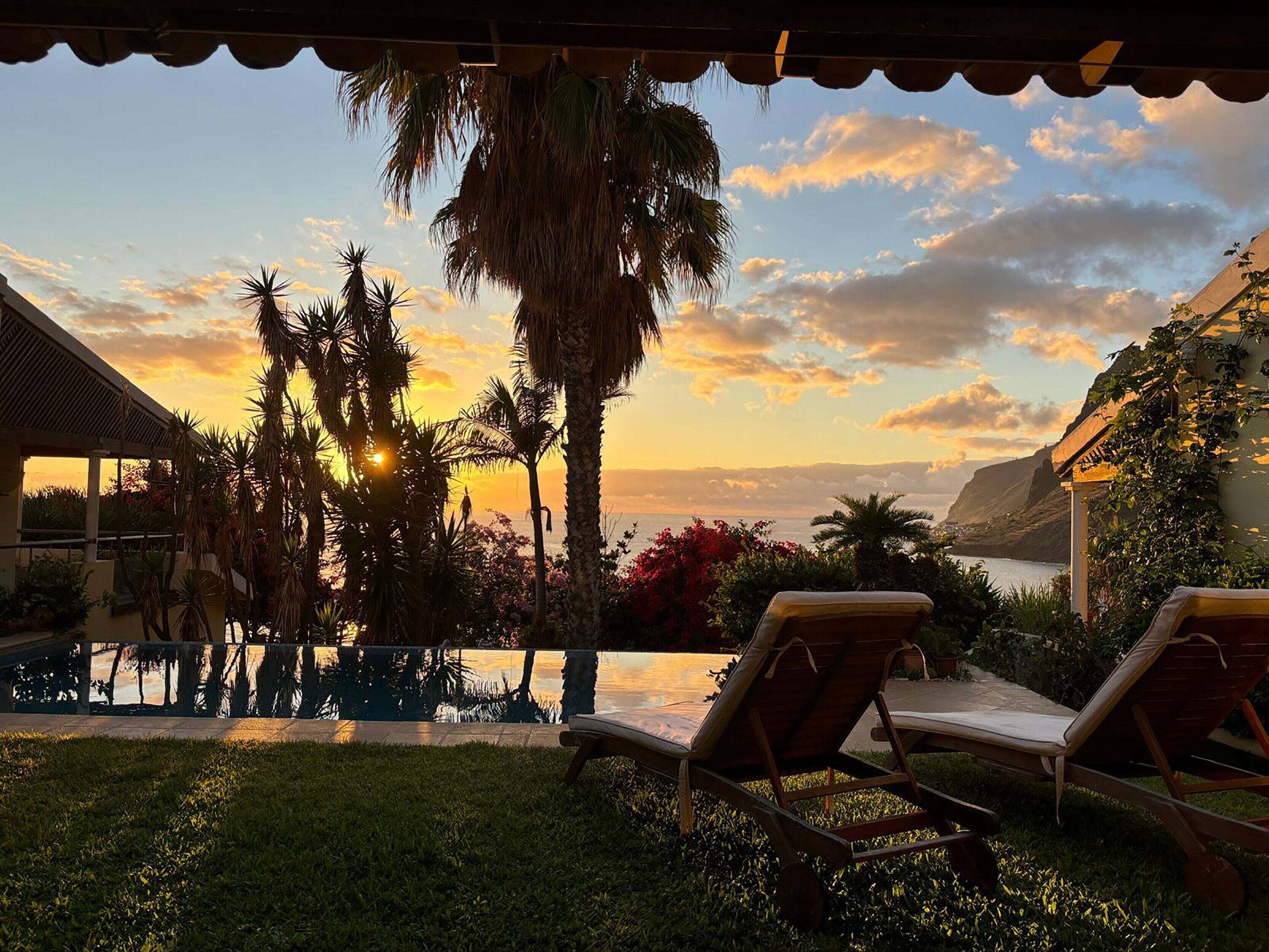 A pool area with lounge chairs, palm trees, and a view of the ocean at sunset.