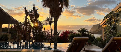 Un área de piscina con sillones de descanso, palmeras y vista al océano al atardecer.