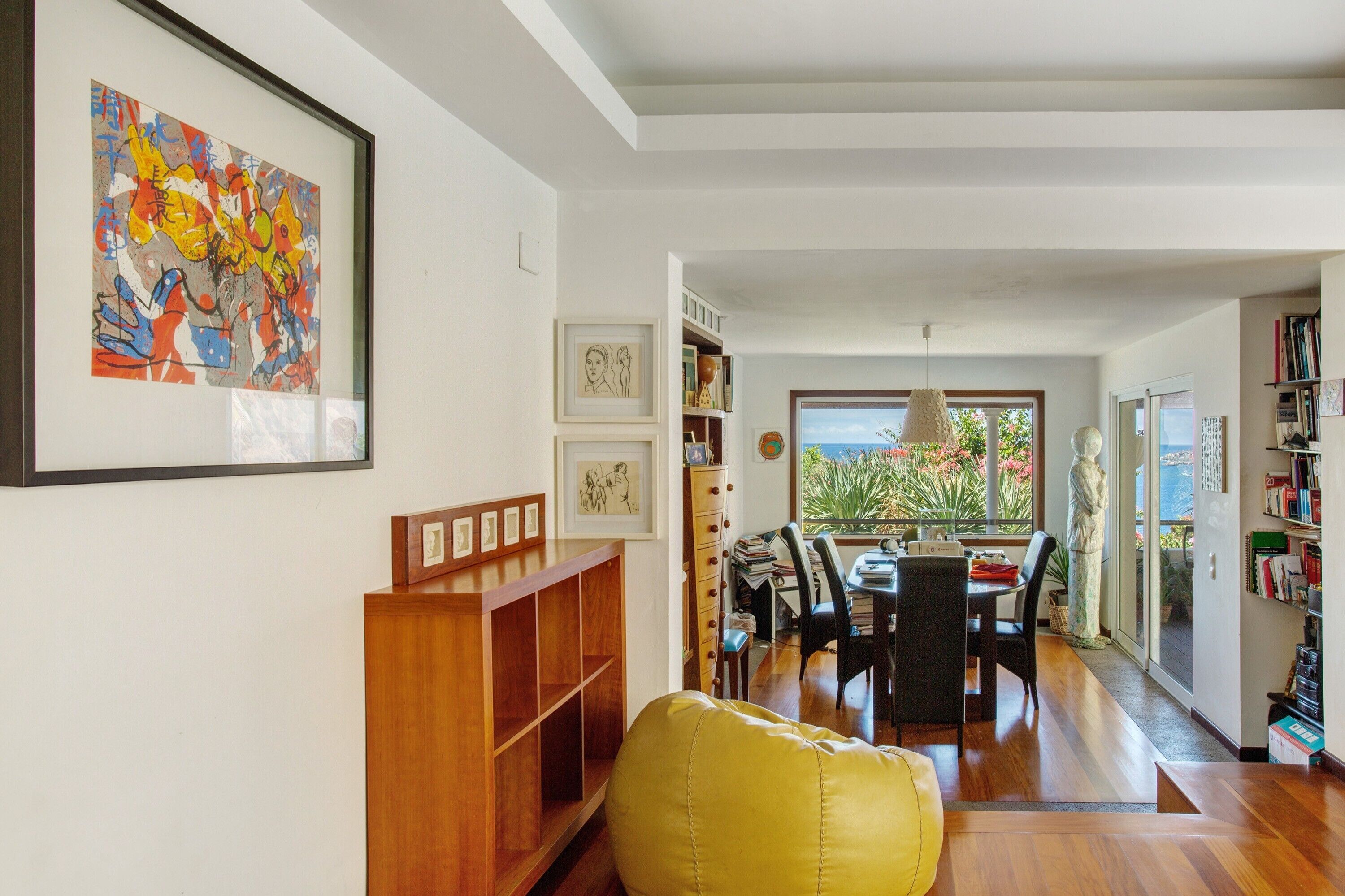 A modern living room with a wooden bookshelf, a yellow armchair, a dining area with a table and chairs, and a view through a glass door to a balcony with plants.
