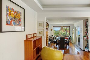 Un salon moderne avec une bibliothèque en bois, un fauteuil jaune, un coin repas avec une table et des chaises, et une vue sur un balcon agrémenté de plantes, accessible par une porte vitrée.