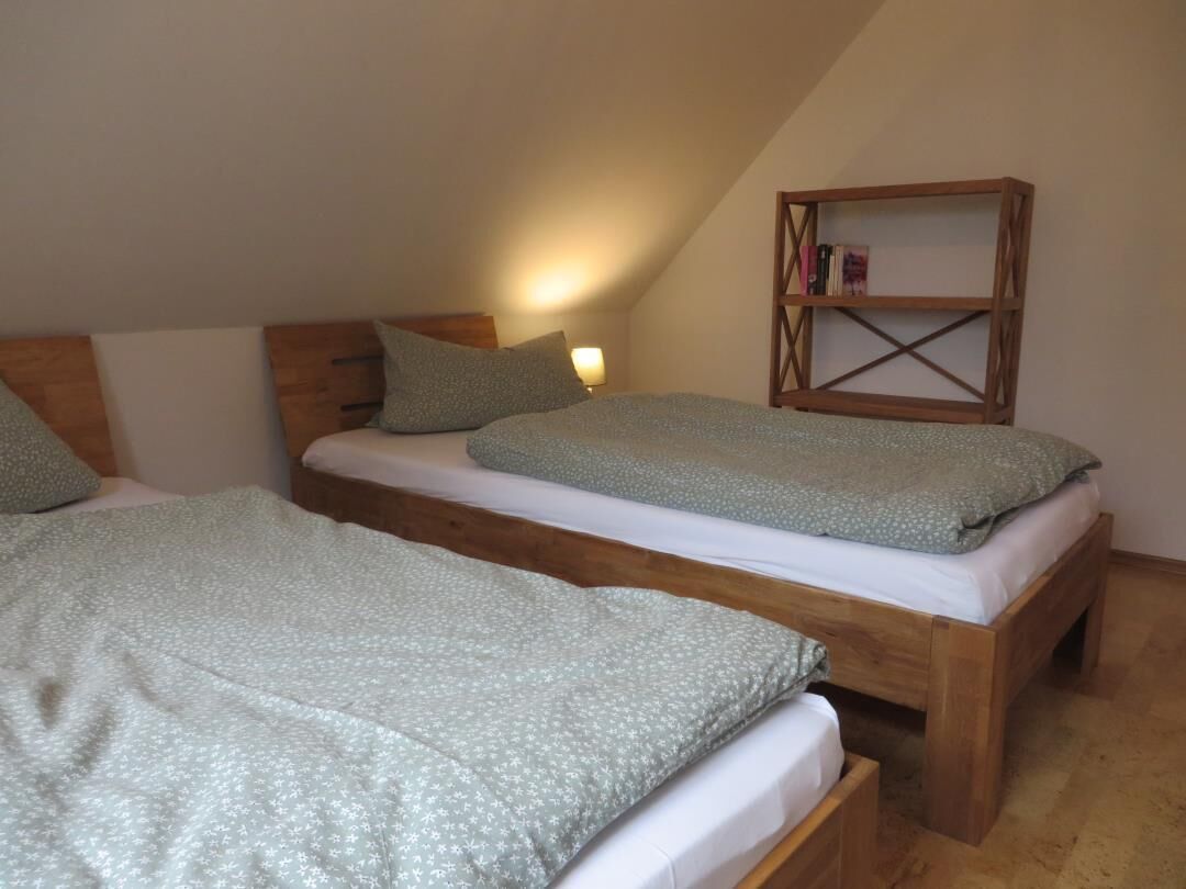 Ein Zimmer mit zwei Holzbettgestellen, jeweils mit einem Kopfkissen und einer Decke. Gegen die Wand steht ein Bücherregal aus Holz.